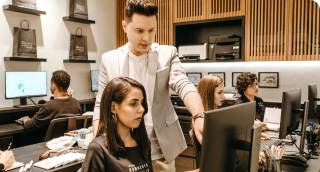 man standing behind woman at desk pointing to monitor